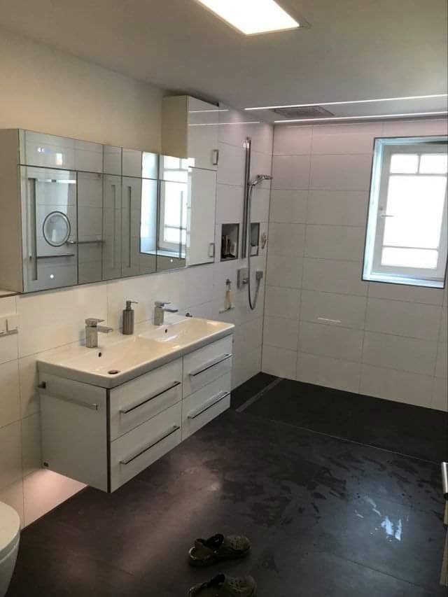 Nowoczesna łazienka z podwójną umywalką na szafce z szufladami, lustrem z szafkami, kabiną prysznicową walk-in z odpływem liniowym i ciemną podłogą, na której leżą klapki.