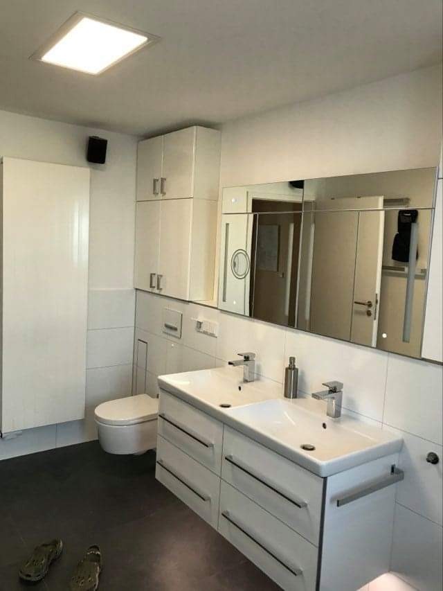 Nowoczesna łazienka z podwójną umywalką, dużym lustrem z oświetleniem LED, białymi szafkami wiszącymi i ciemną podłogą. W tle widoczna toaleta i biały grzejnik. Na podłodze leżą klapki.