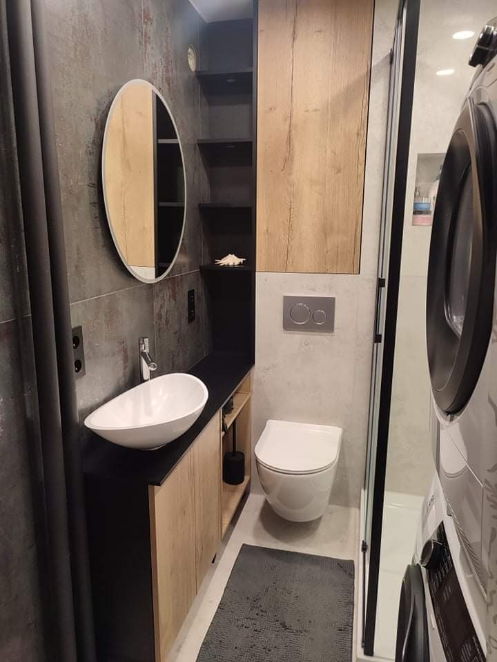 Nowoczesna, wąska łazienka z umywalką nablatową, toaletą podwieszaną, zabudową z drewnopodobnym frontem i pralką za szklaną kabiną prysznicową.