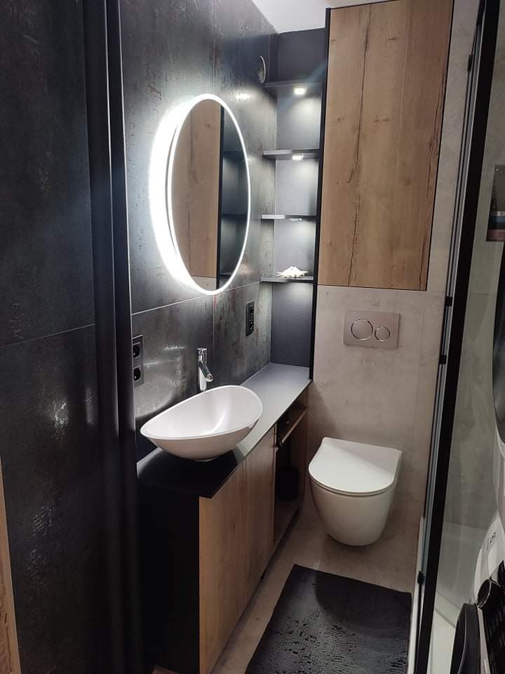 Nowoczesna łazienka z owalną umywalką nablatową, podświetlanym okrągłym lustrem, wiszącą toaletą i szafką z drewnopodobnym frontem, wykończona w ciemnych szarościach i jasnych beżach.