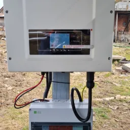 Biały inwerter marki Sofar Solar zamontowany na słupku, z widocznym panelem sterowania i skrzynką elektryczną poniżej, na tle trawy i fragmentu budynku.