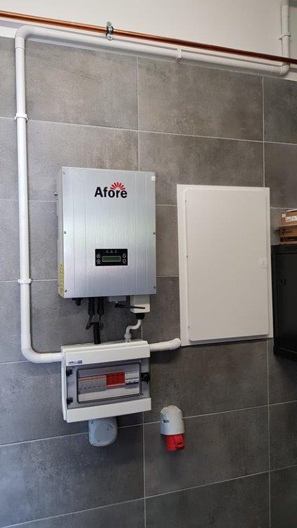 Srebrny falownik z logo Afore zamontowany na szarej, kafelkowanej ścianie, z widoczną skrzynką bezpiecznikową i gniazdem siłowym, oraz białymi rurami instalacyjnymi.