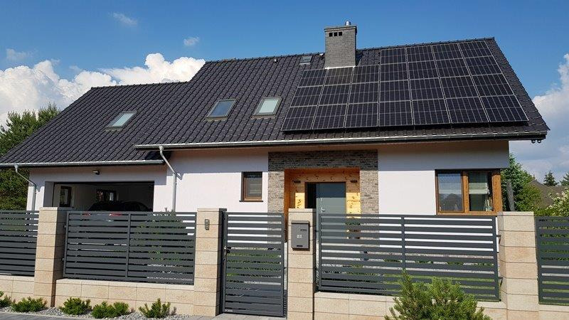 Nowoczesny dom jednorodzinny z ciemną dachówką i panelami słonecznymi na dachu, widoczne okna dachowe, szary płot lamelowy z kamiennymi słupkami i skrzynką na listy.