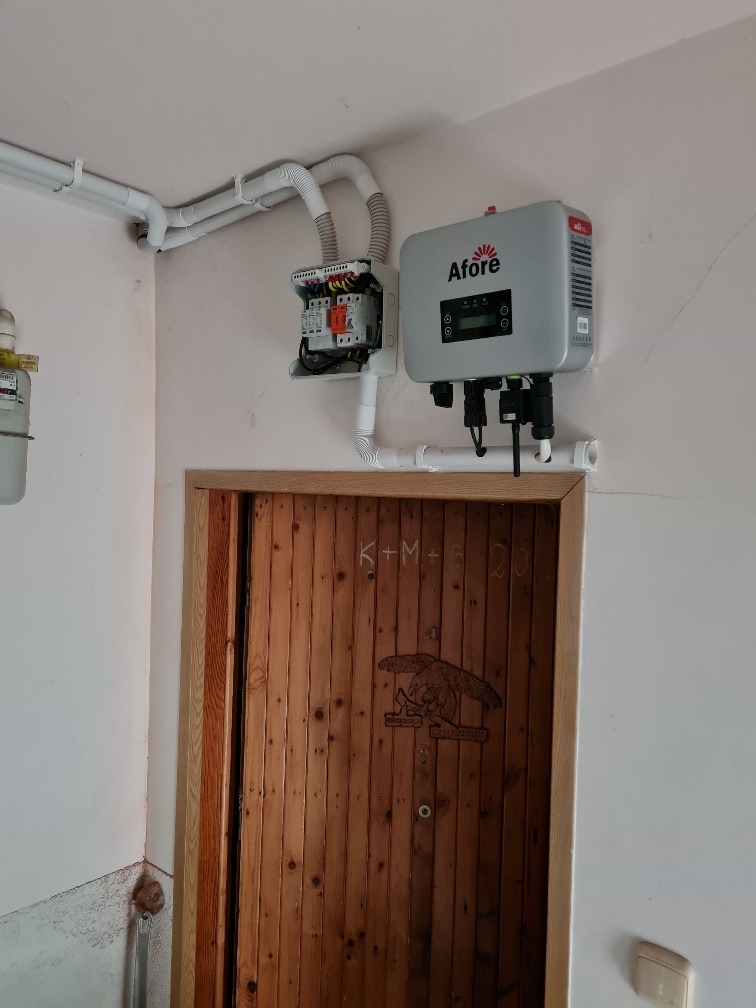 Szary inwerter marki Afore zamontowany na ścianie nad drewnianymi drzwiami z wyrytym orłem, połączony rurami instalacyjnymi z rozdzielnicą elektryczną.