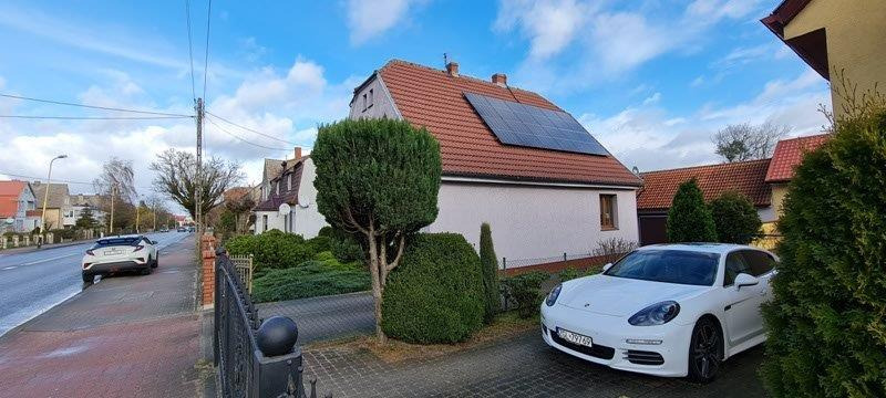 Dom jednorodzinny z czerwoną dachówką i panelami słonecznymi na dachu, biały samochód zaparkowany na podjeździe, bujna zieleń w ogrodzie.