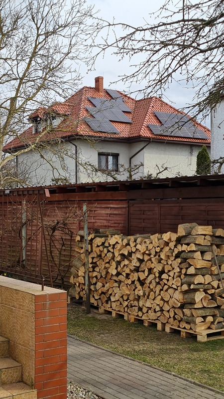 Dom jednorodzinny z czerwoną dachówką, na której zamontowano panele słoneczne; w tle widoczne drzewa bez liści i sterta drewna kominkowego.