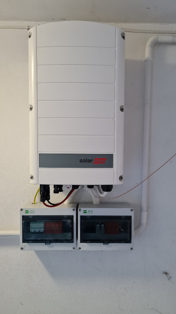 Biały falownik SolarEdge zamontowany na ścianie, pod nim dwie białe skrzynki z bezpiecznikami, widoczne kable i rury instalacyjne.