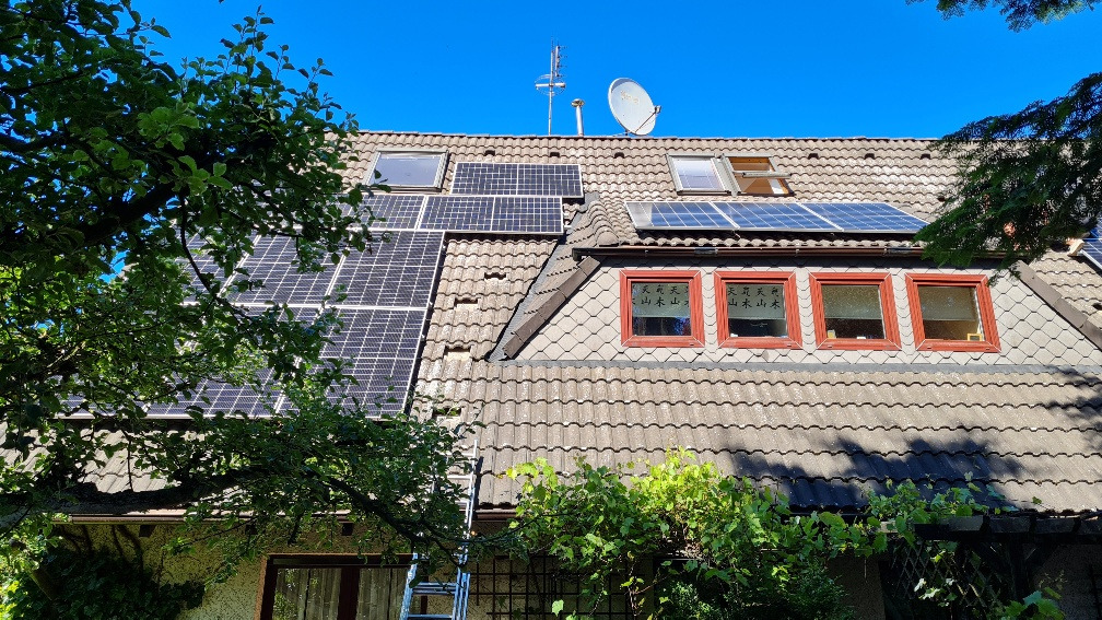 Instalacja paneli słonecznych na dachu domu z dachówką, widoczne okna dachowe, antena satelitarna i drabina oparta o ścianę, częściowo zasłonięte przez gałęzie drzew.