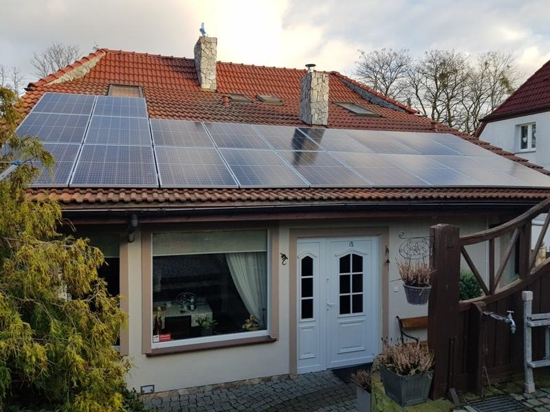 Dom jednorodzinny z panelami słonecznymi na dachu z czerwonej dachówki, widok z poziomu chodnika, na pierwszym planie brama i rośliny w donicach.