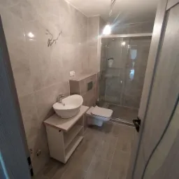 Wykończona łazienka w odcieniach szarości z kabiną prysznicową, umywalką nablatową na szafce z półkami, wiszącą toaletą i ozdobnymi haczykami na ścianie.