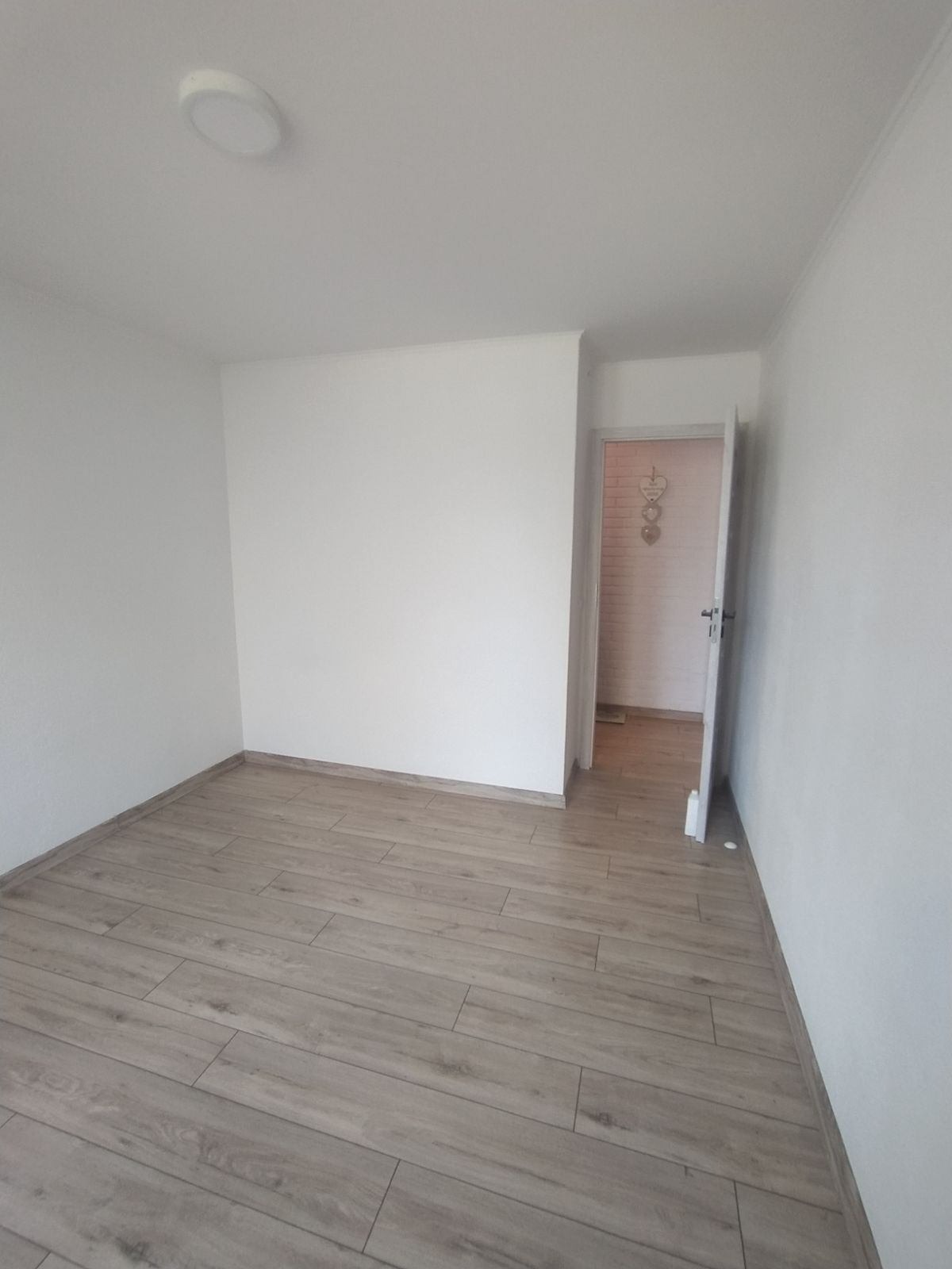 Puste, odnowione wnętrze z panelami podłogowymi imitującymi drewno i białymi ścianami, widok na otwarte drzwi do łazienki z dekoracją na ścianie.