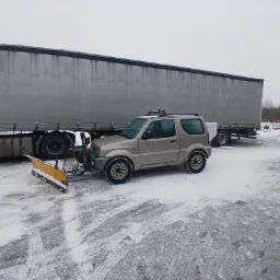Beżowy samochód terenowy z żółtym pługiem do śniegu na parkingu pokrytym śniegiem, obok zaparkowana ciężarówka z naczepą.