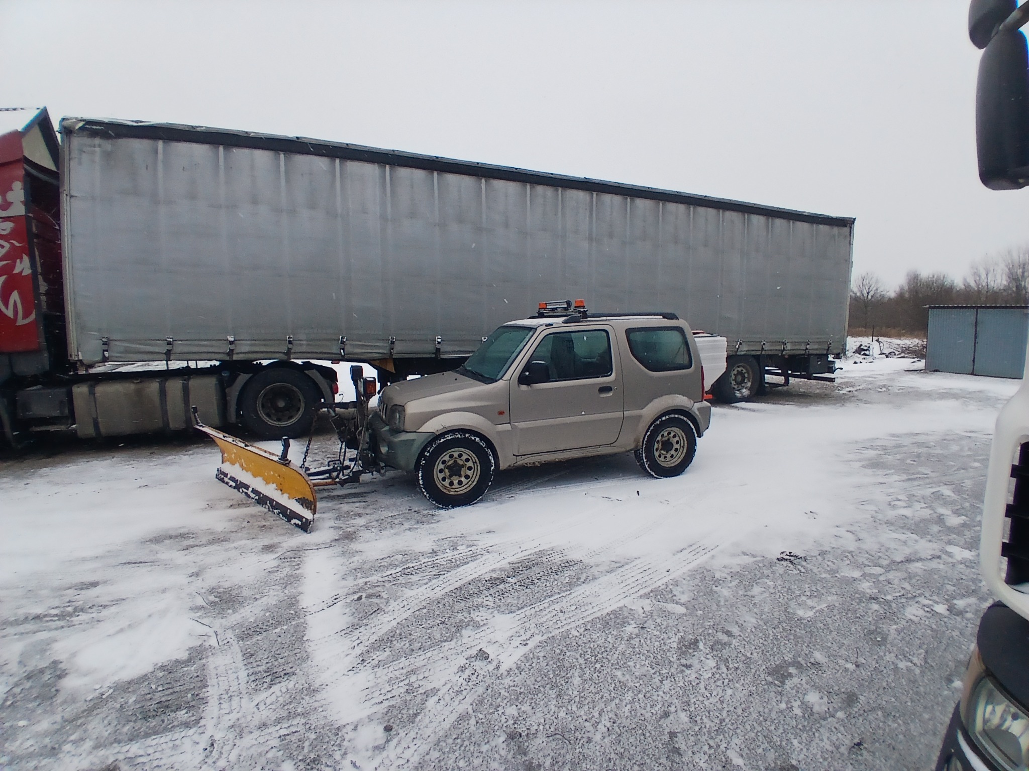 Beżowy samochód terenowy z żółtym pługiem do śniegu na parkingu pokrytym śniegiem, obok zaparkowana ciężarówka z naczepą.