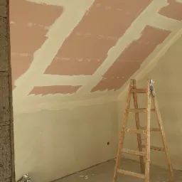 Poddasze w trakcie wykańczania: widoczne płyty kartonowo-gipsowe na skosach sufitu, częściowo zaszpachlowane, drewniana drabina w rogu, worek z zaprawą na podłodze.