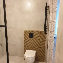 Nowoczesna łazienka z beżowymi płytkami, wiszącą toaletą na tle dekoracyjnej ściany z drewnianymi wzorami w romby i czarnym przyciskiem spłukiwania, oraz czarnym grzejnikiem drabinkowym.