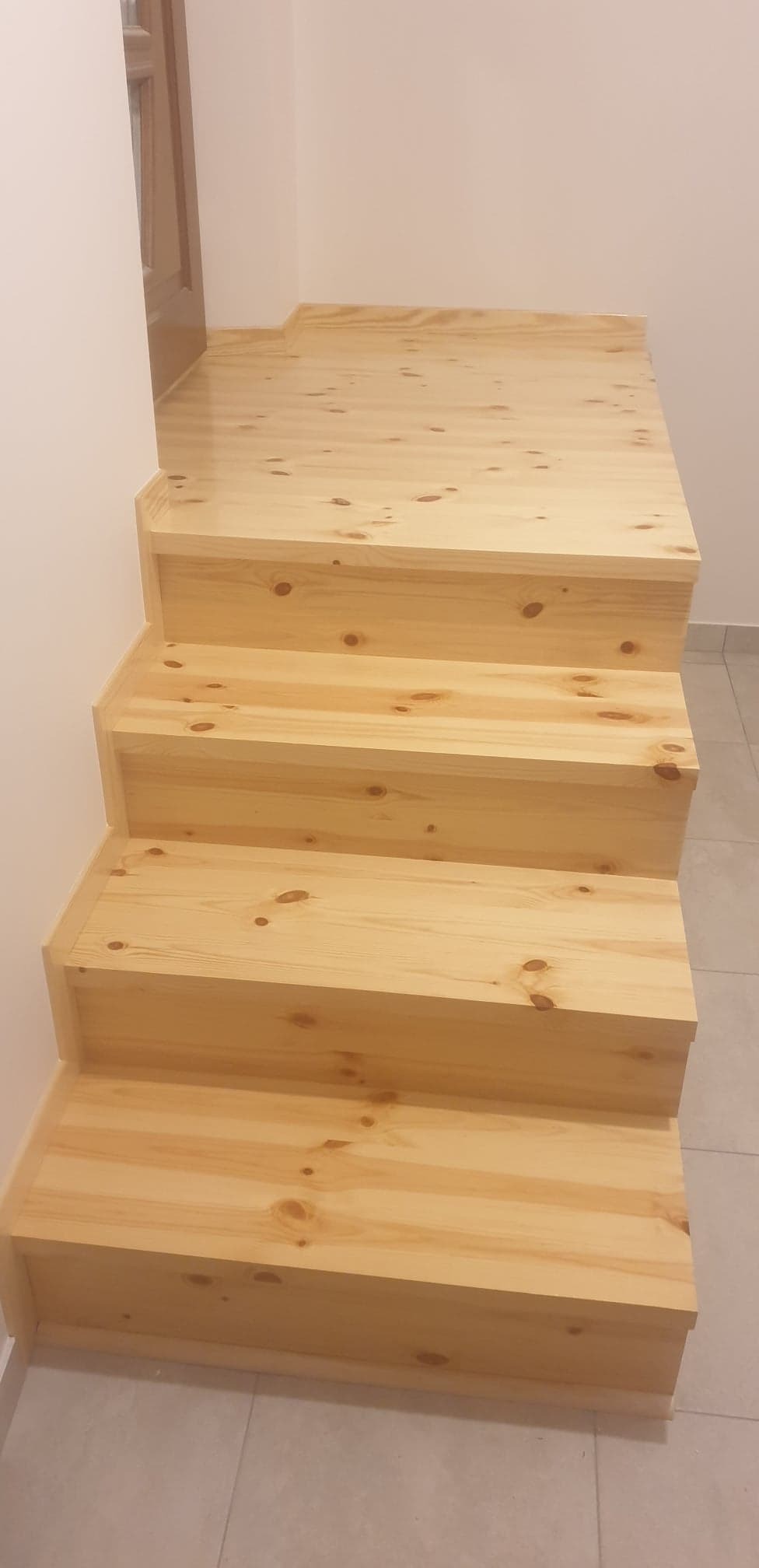 Nowe, drewniane schody sosnowe z widocznymi słojami, prowadzące na podest, przy białej ścianie i szarej podłodze.