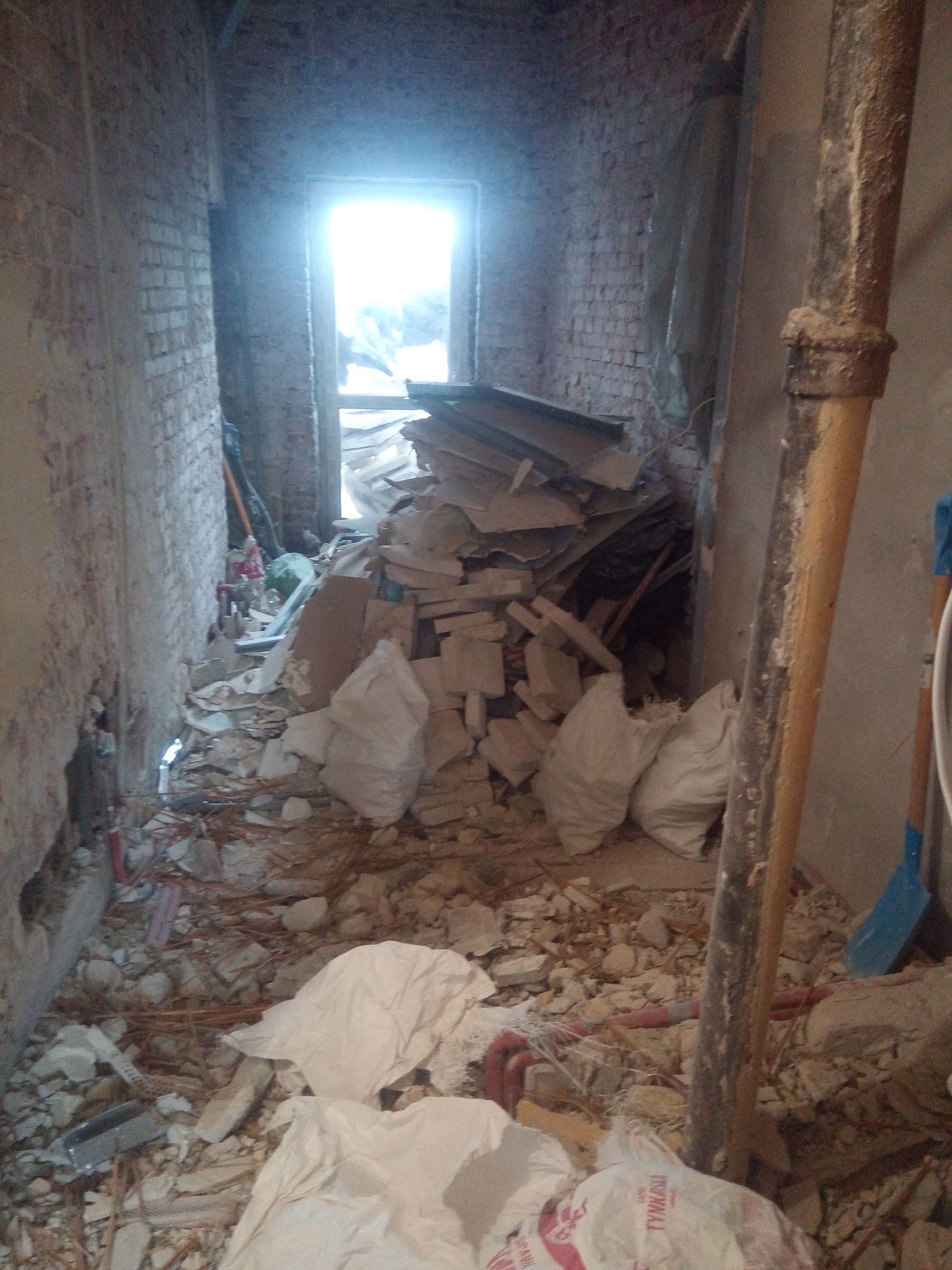 Korytarz w trakcie demontażu, widoczne gruz, worki z odpadami budowlanymi, odsłonięta cegła, rury i narzędzia, z widokiem na jasne światło zza otworu drzwiowego.
