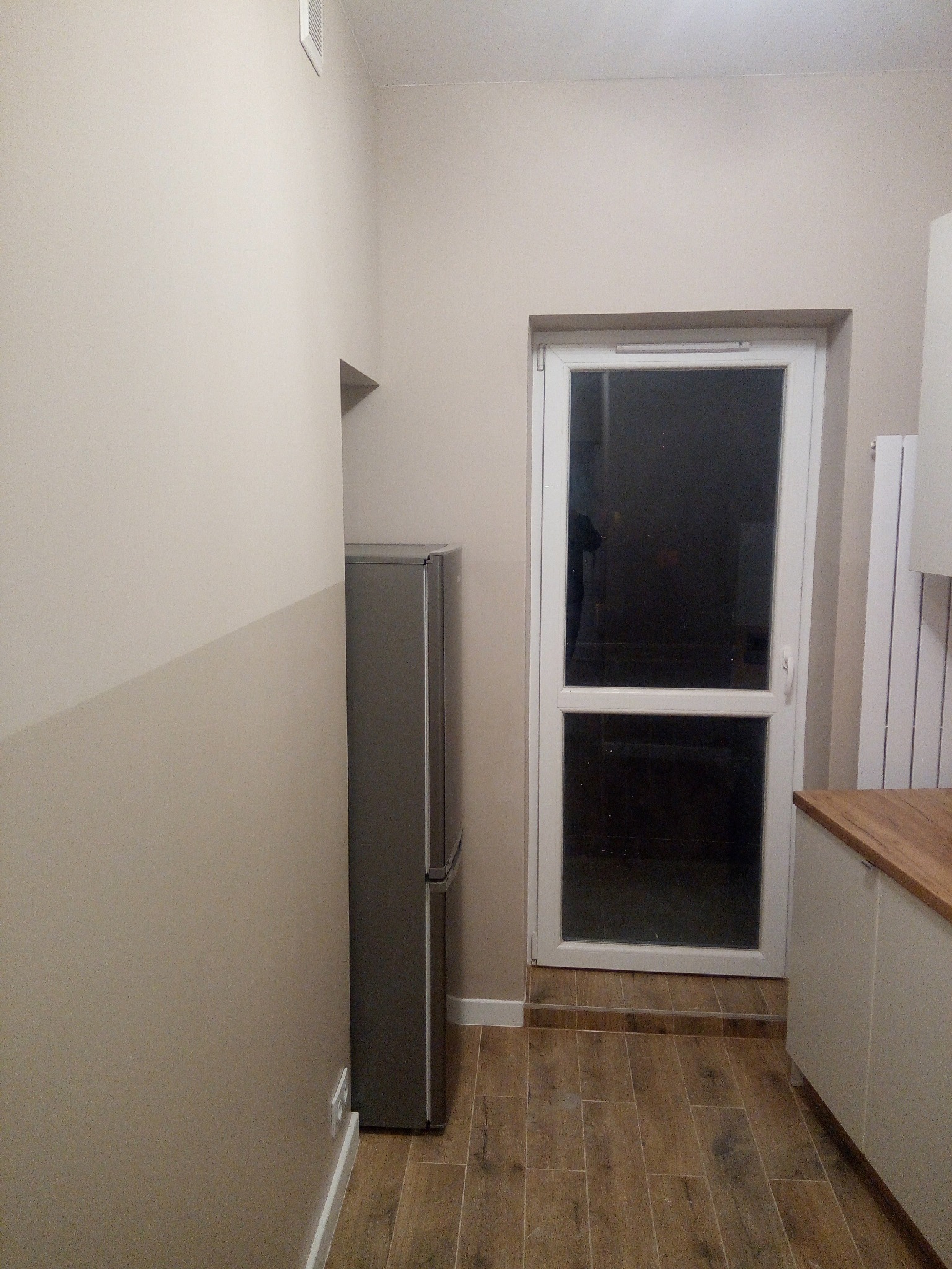 Kuchnia po remoncie: beżowe ściany z szarym pasem w dolnej części, drewnopodobne płytki podłogowe, białe meble kuchenne z drewnianym blatem, szara lodówka i białe drzwi balkonowe.