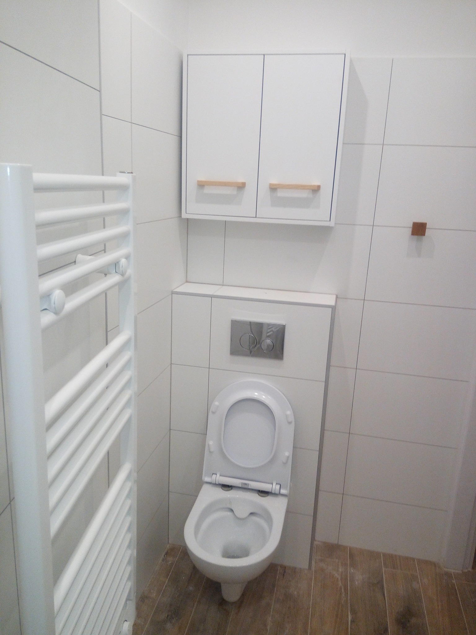 Biała, nowoczesna łazienka z wiszącą szafką nad toaletą, grzejnikiem drabinkowym i podłogą imitującą drewno, wykończona białymi, kwadratowymi płytkami z szarą fugą.
