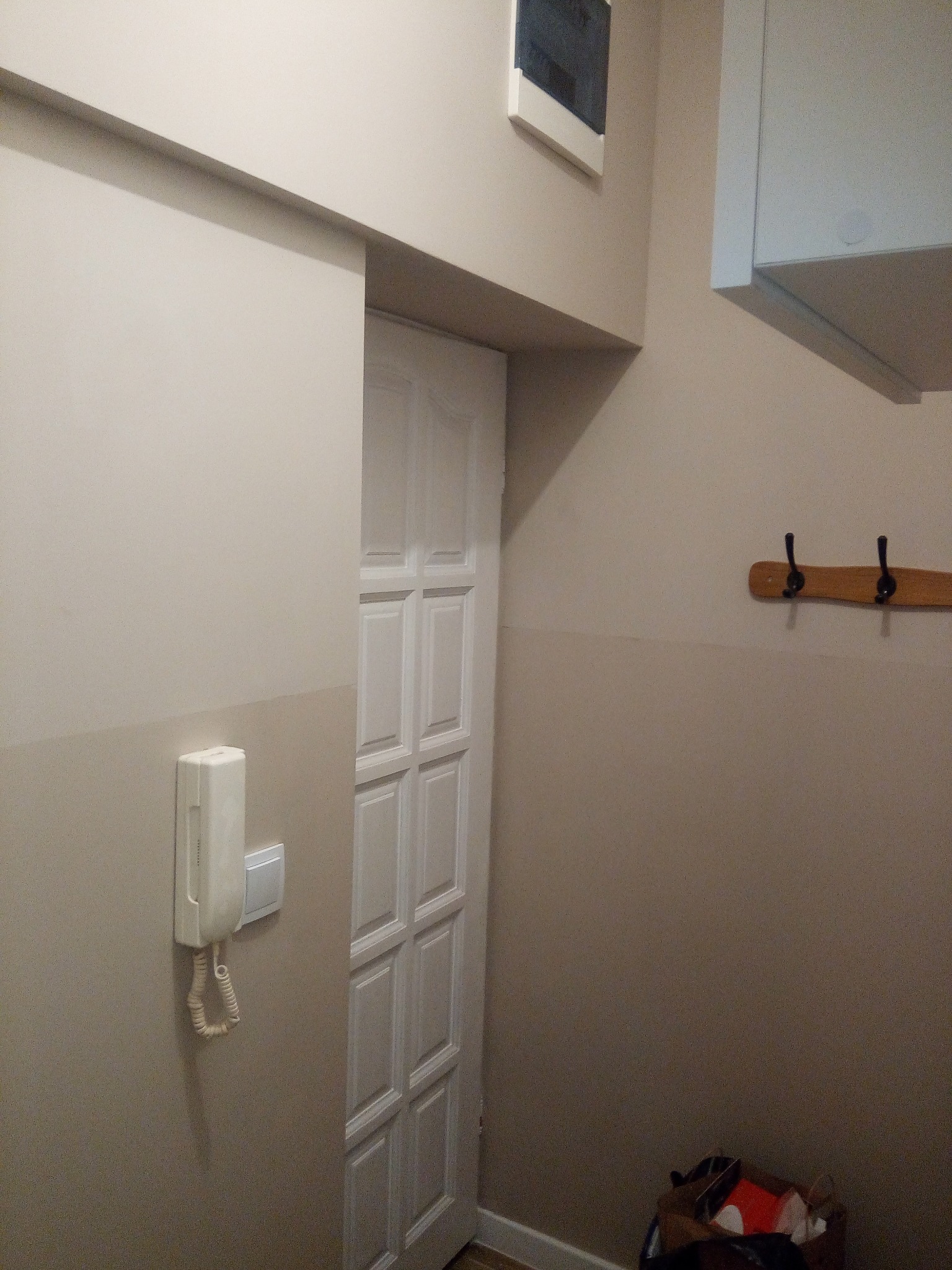 Korytarz po remoncie z pomalowanymi ścianami na dwa odcienie beżu, białe drzwi z panelami, wiszący telefon na ścianie i wieszak z haczykami.