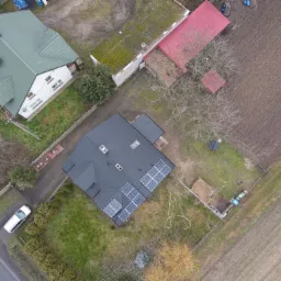 Widok z drona na dom z ciemnym dachem i panelami słonecznymi, otoczony zielenią i polami, z widocznym samochodem na drodze.