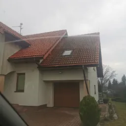 Dom jednorodzinny z dwuspadowym dachem z czerwonej dachówki, z widocznym fragmentem dachu z jednym oknem dachowym, elewacja w kolorze jasnego beżu, rynny brązowe, na podjeździe kostka brukowa,...