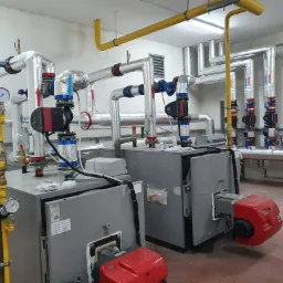 Dwa szare kotły grzewcze z czerwonymi palnikami, rury w izolacji z oznaczeniami przepływu, manometry oraz żółta instalacja gazowa w kotłowni.