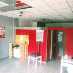 Pakersi Rumia- Usługi Kurierskie - Wnętrze punktu obsługi przesyłek: waga elektroniczna, cennik na ścianie, regał z kartonami, stanowisko do pakowania z palet, plakat reklamowy 'Pakersi'.