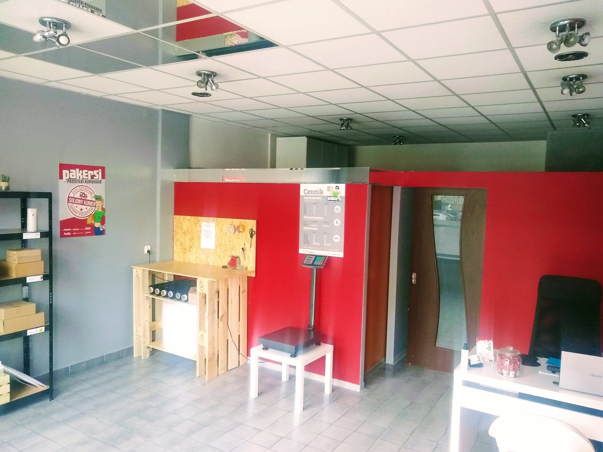 Wnętrze punktu obsługi przesyłek: waga elektroniczna, cennik na ścianie, regał z kartonami, stanowisko do pakowania z palet, plakat reklamowy 'Pakersi'.