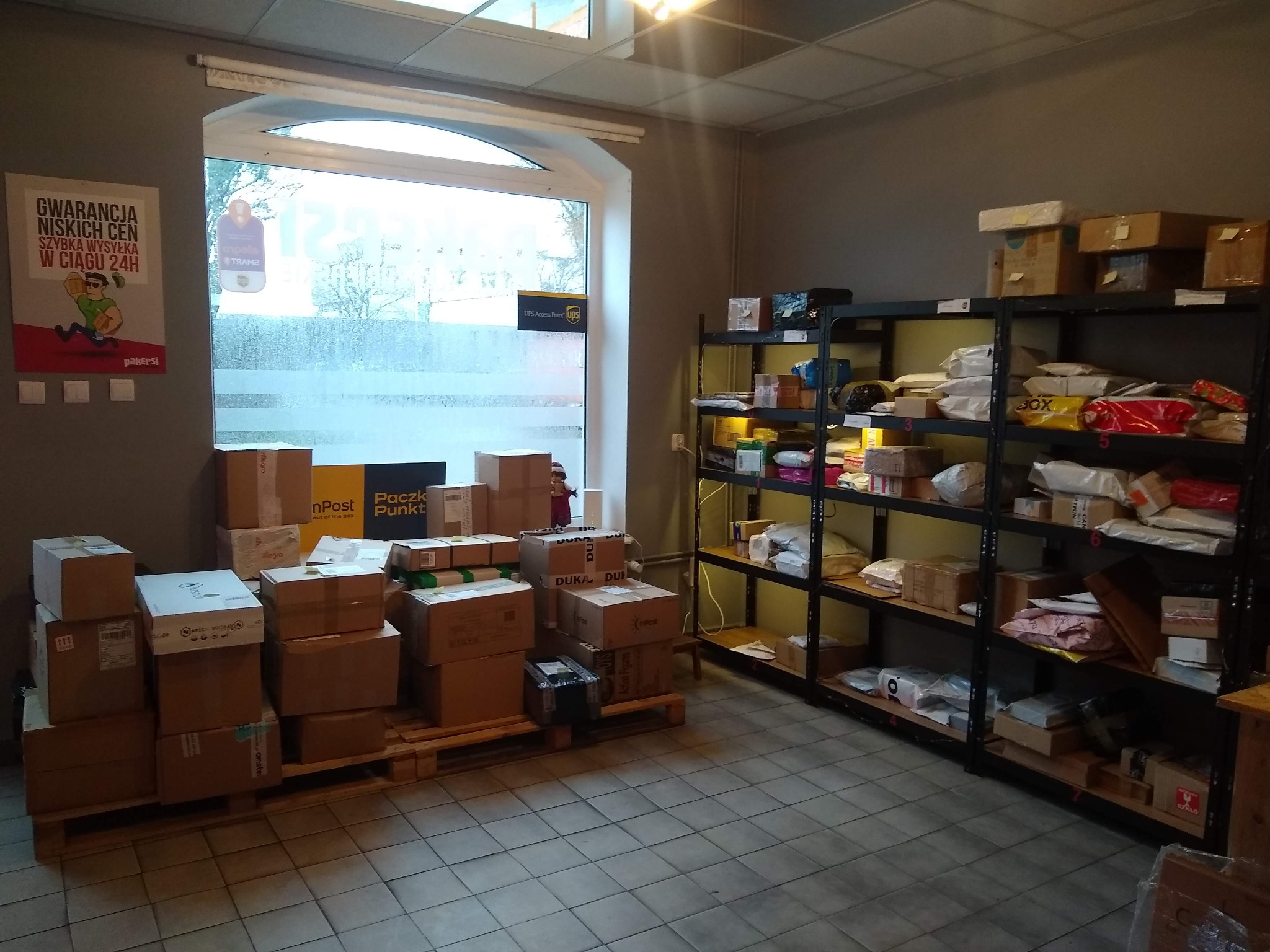 Wnętrze punktu kurierskiego z paczkami ułożonymi na palecie i na metalowych regałach, widoczne logo UPS Access Point i reklama firmy kurierskiej na ścianie.