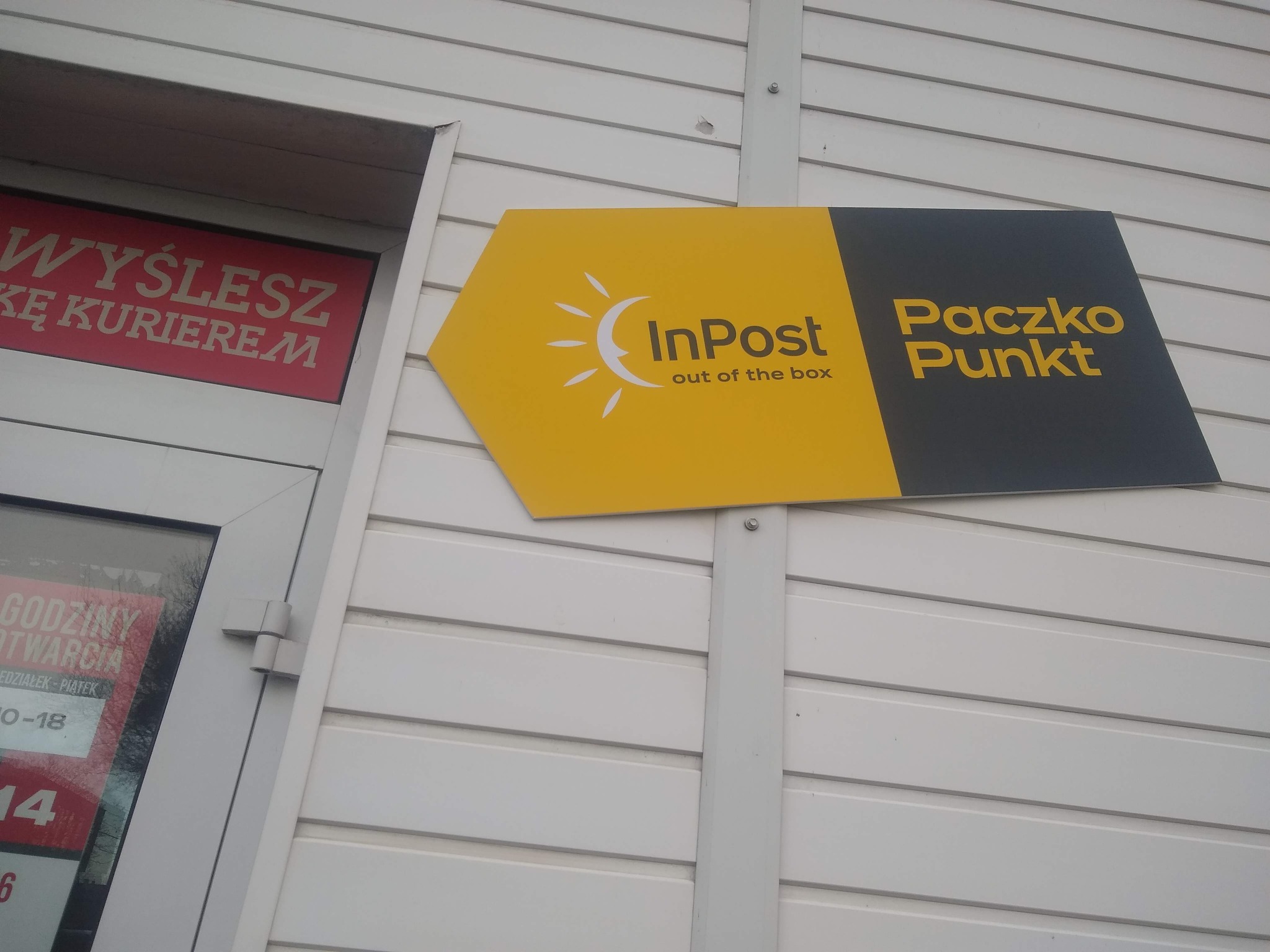 Żółto-czarny szyld InPost Paczko Punkt zamontowany na białej ścianie budynku, widoczny fragment plakatu z napisem 'Wyślesz Kę Kurierem'.