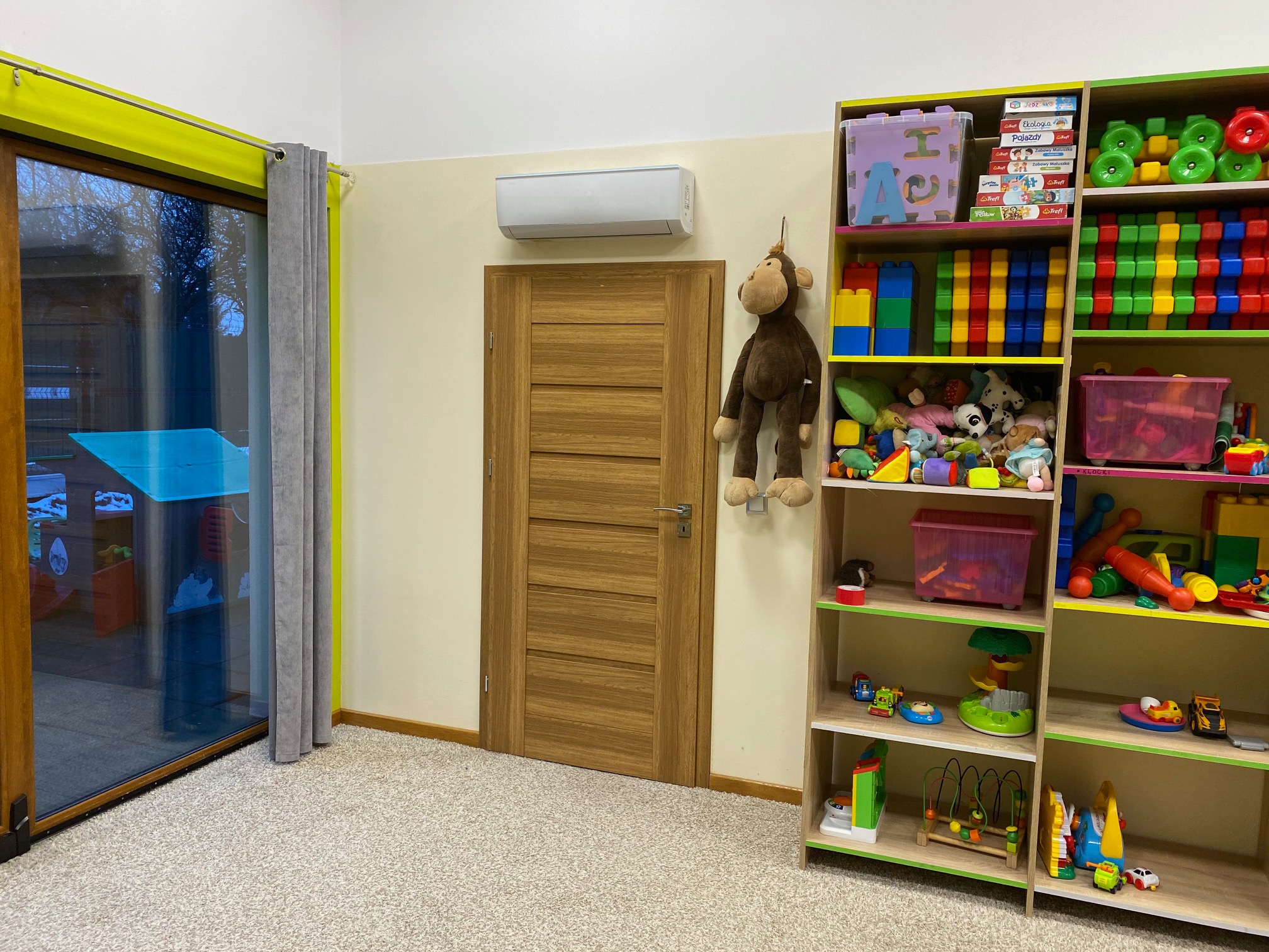 Wnętrze pokoju dziecięcego z zamontowanym klimatyzatorem nad drewnianymi drzwiami, obok okno z zieloną ramą i regał z zabawkami.