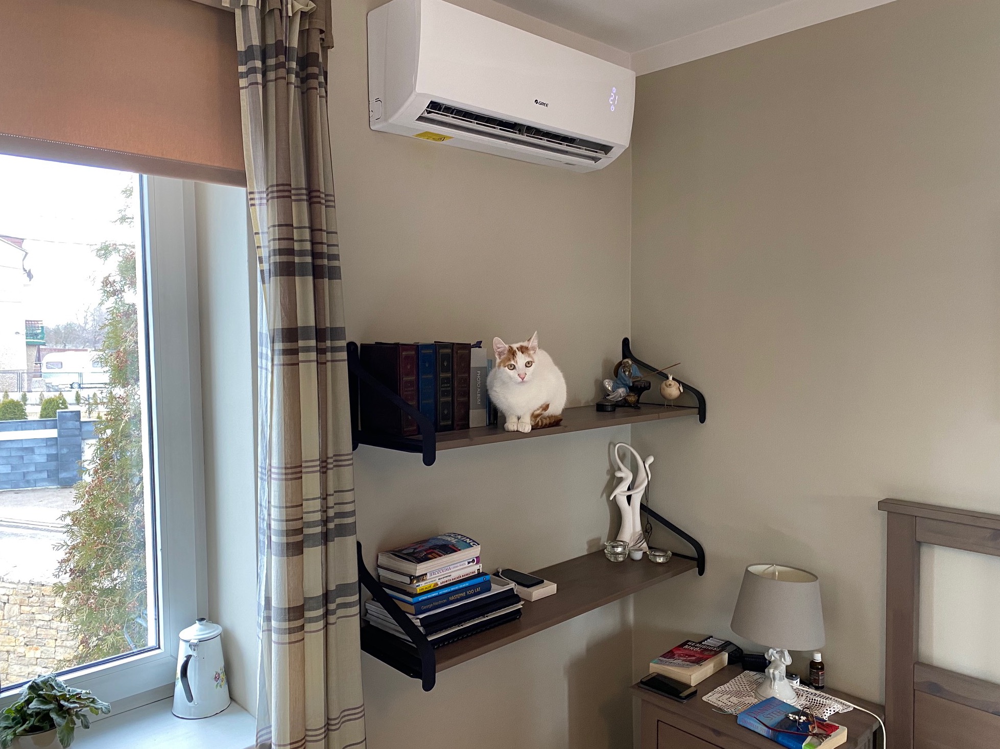 Biały klimatyzator z wyświetlaczem temperatury zamontowany nad półkami z książkami, na których siedzi biało-rudy kot w pokoju z oknem i zasłoną.