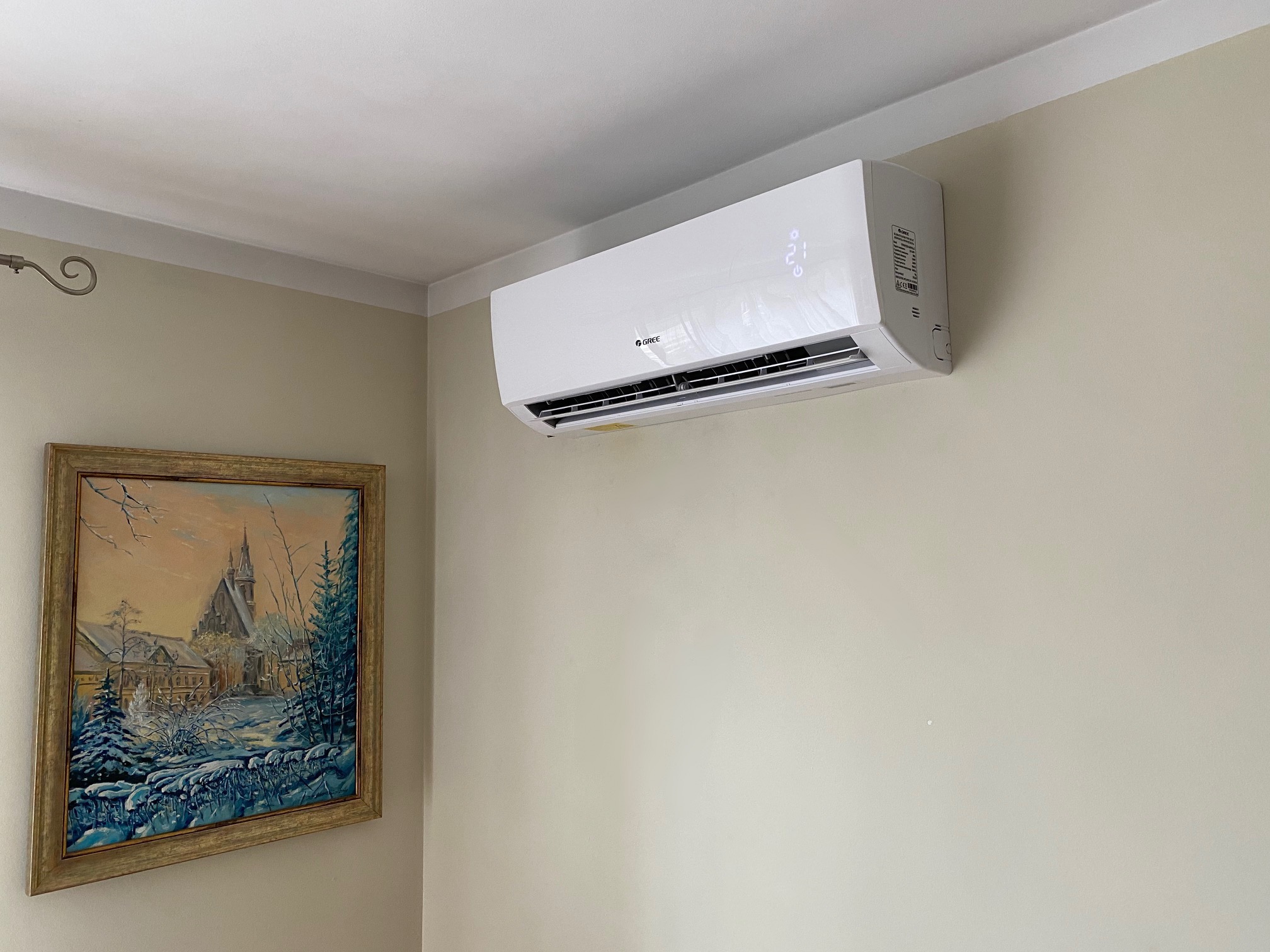 Biała klimatyzacja marki Gree zamontowana na ścianie w pokoju z beżowymi ścianami, obok wisi obraz w złotej ramie przedstawiający zimowy krajobraz.