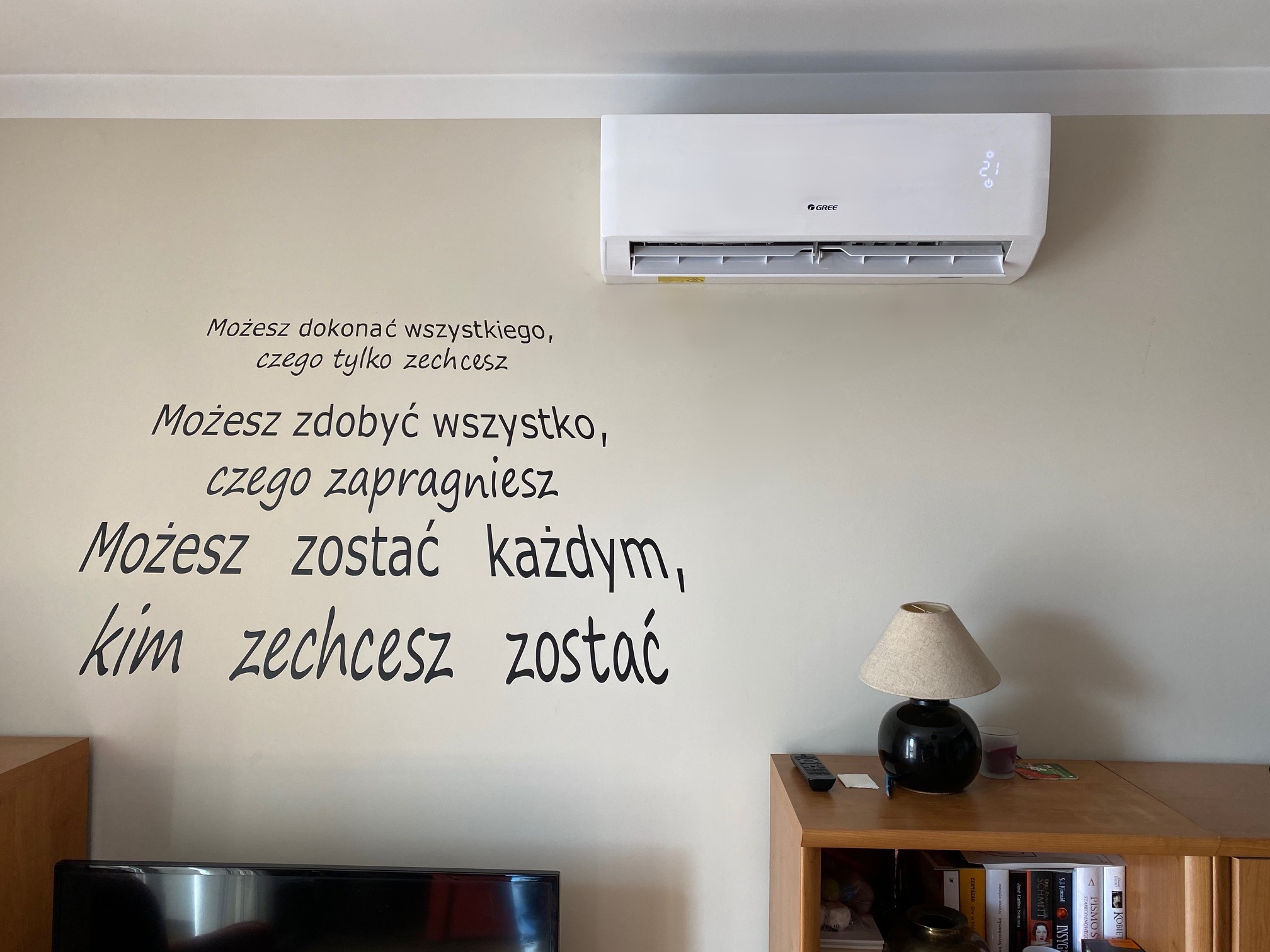 Biały klimatyzator marki Gree zamontowany na ścianie z naklejką motywacyjną w salonie z meblami.