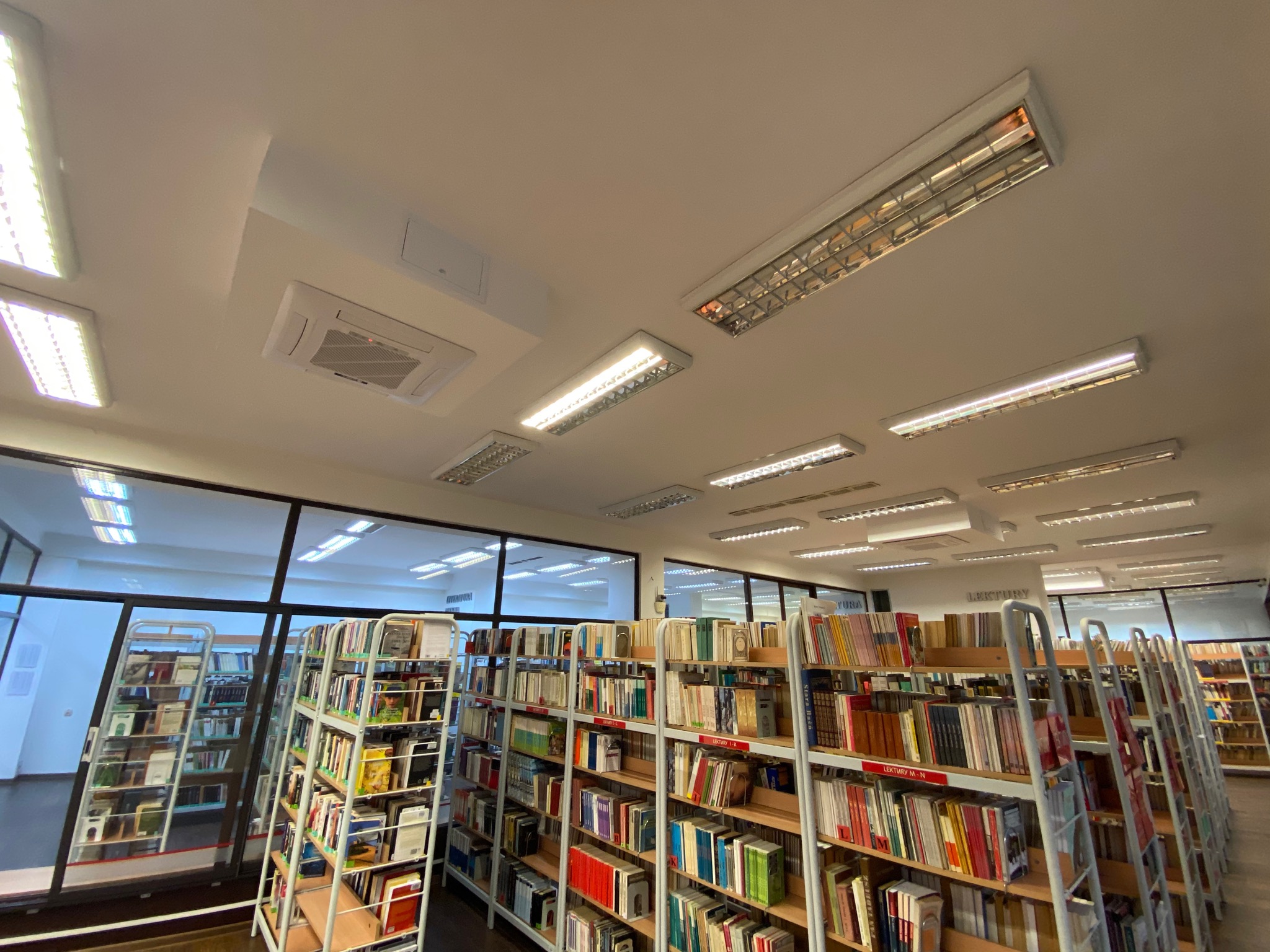 Kasetonowy klimatyzator widoczny na suficie biblioteki w Olkuszu, pomiędzy rzędami świetlówek i regałami z książkami.