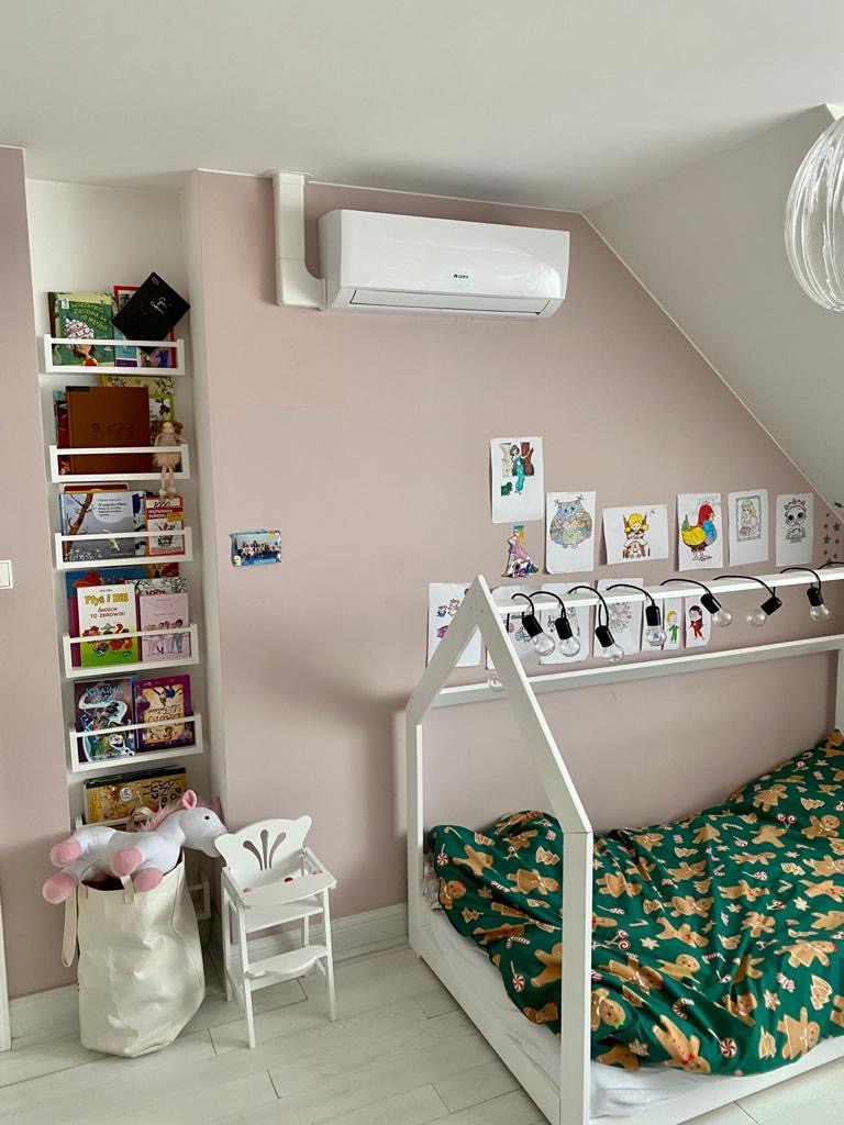Biały klimatyzator ścienny w pokoju dziecięcym z różowymi ścianami, książkami i pluszowym jednorożcem.