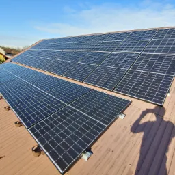 Instalacja paneli fotowoltaicznych na brązowym blaszanym dachu domu jednorodzinnego w słoneczny dzień, widoczny pracownik z narzędziami na dachu.