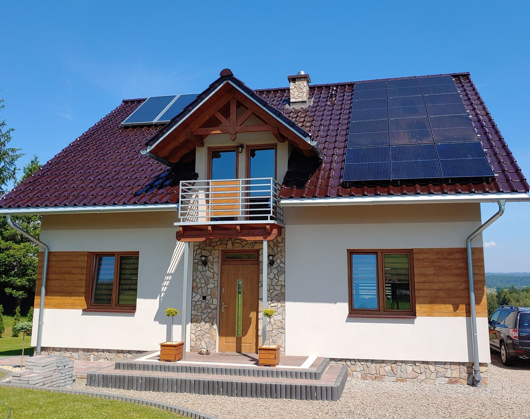 Dom jednorodzinny z brązową dachówką i panelami słonecznymi na dachu, balkon z metalową barierką, elewacja z elementami kamiennymi i drewnianymi, widok z przodu w słoneczny dzień.