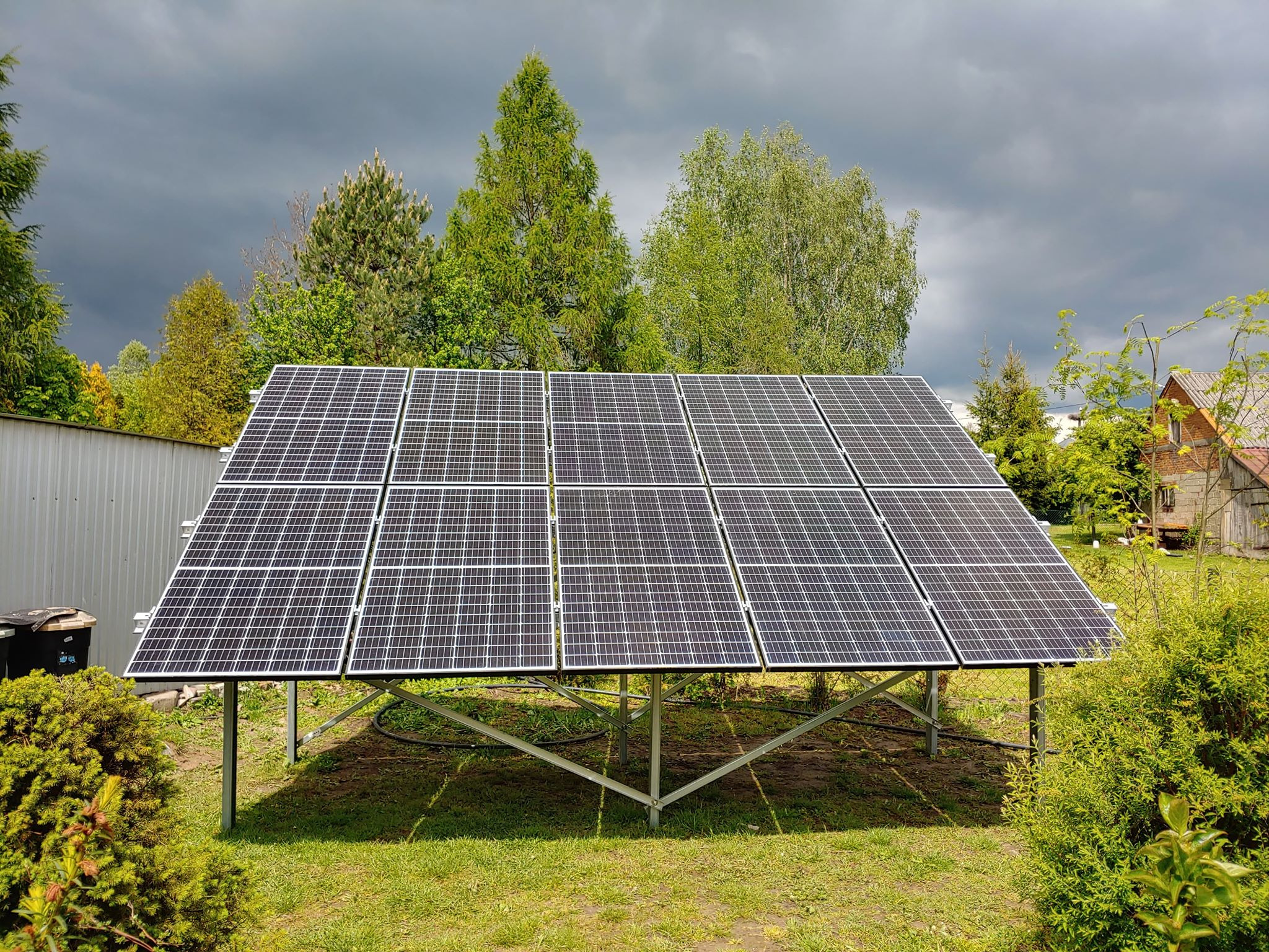 Panel fotowoltaiczny na metalowej konstrukcji, umieszczony na trawniku przed budynkiem gospodarczym i domem, na tle pochmurnego nieba.