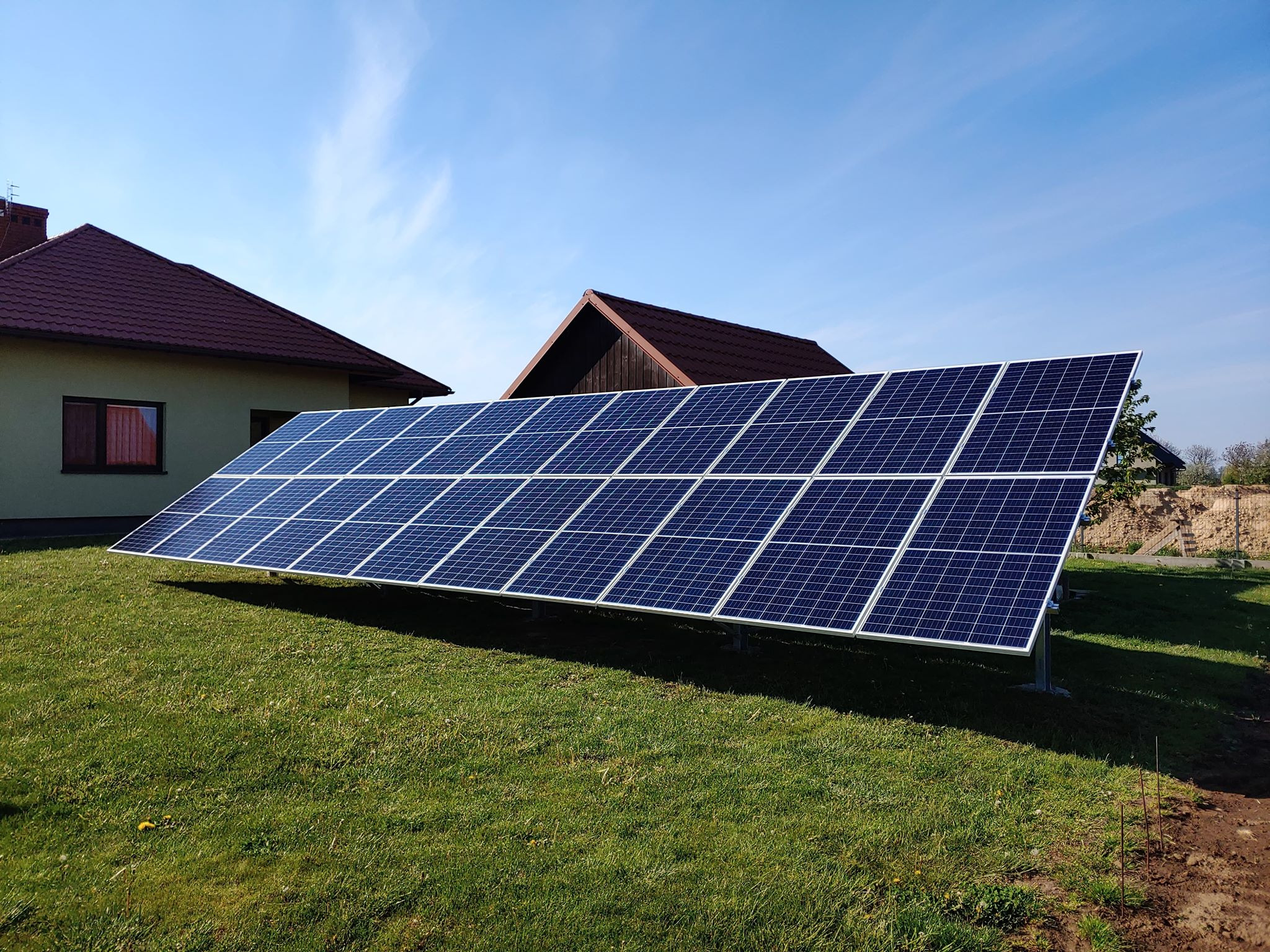Panel fotowoltaiczny zamontowany na metalowych podporach na trawniku przed domem z czerwoną dachówką, widok w słoneczny dzień.