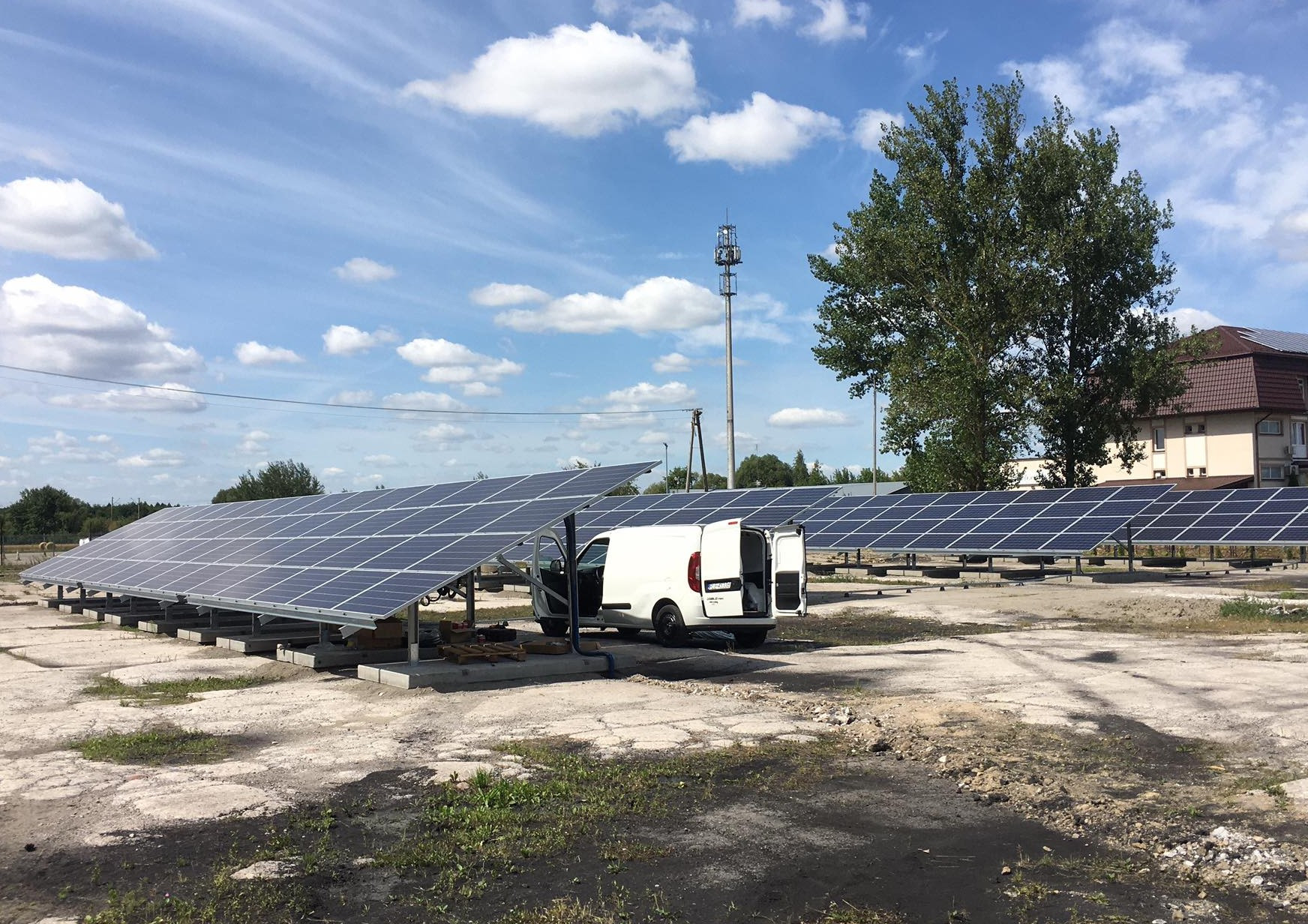 Rząd paneli słonecznych na betonowych podstawach, biały samochód dostawczy z otwartymi drzwiami w tle, błękitne niebo z chmurami, widoczna wieża telekomunikacyjna w oddali.