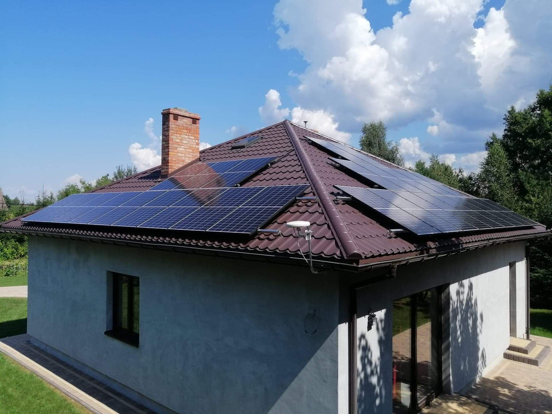 Dom jednorodzinny z brązowym dachem pokrytym panelami fotowoltaicznymi, widoczny komin z cegły i biała antena satelitarna na dachu, błękitne niebo z białymi chmurami w tle.