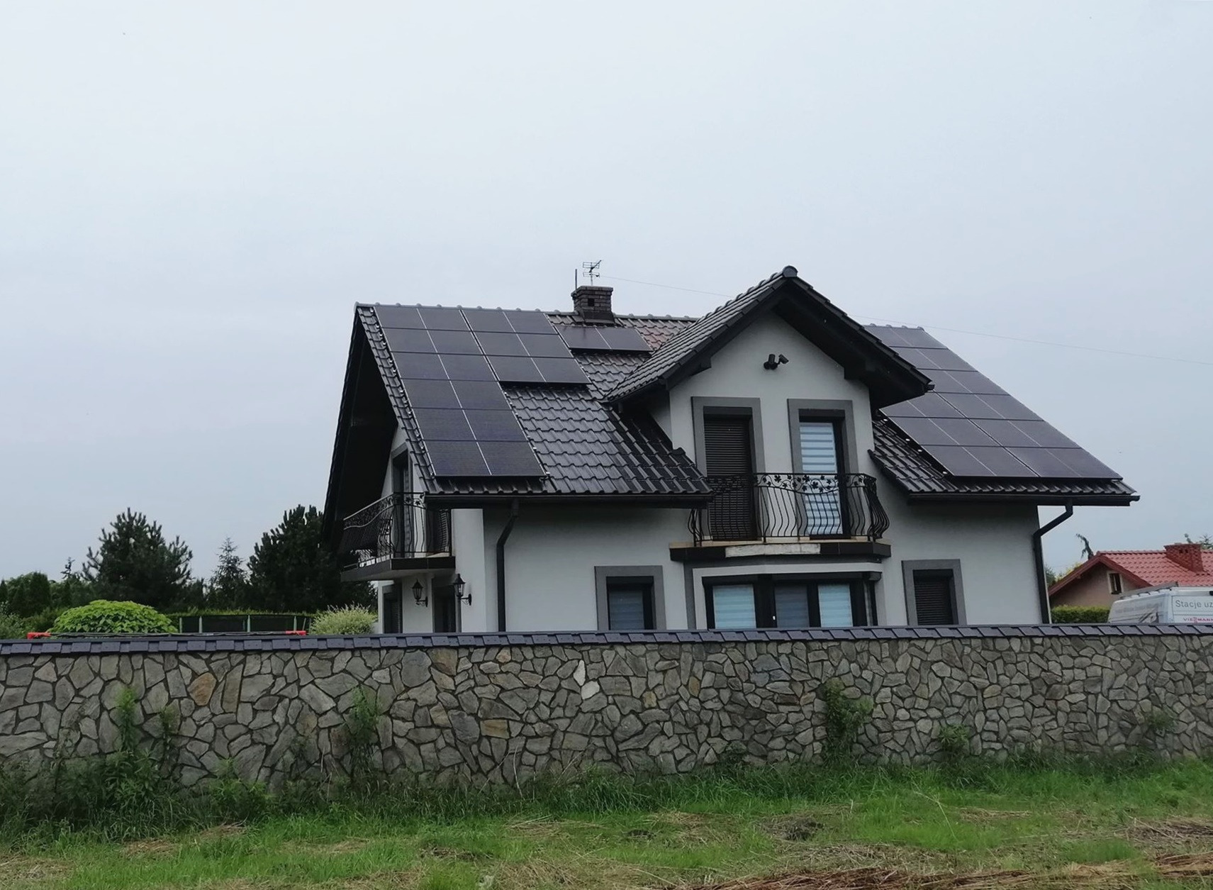 Dom jednorodzinny z zainstalowanymi panelami fotowoltaicznymi na dachu, widok zza kamiennego ogrodzenia.