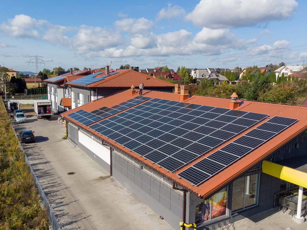 Widok z góry na budynek z pomarańczowym dachem pokrytym panelami fotowoltaicznymi, na dachu stoi człowiek, w tle zabudowa jednorodzinna i błękitne niebo z białymi chmurami.