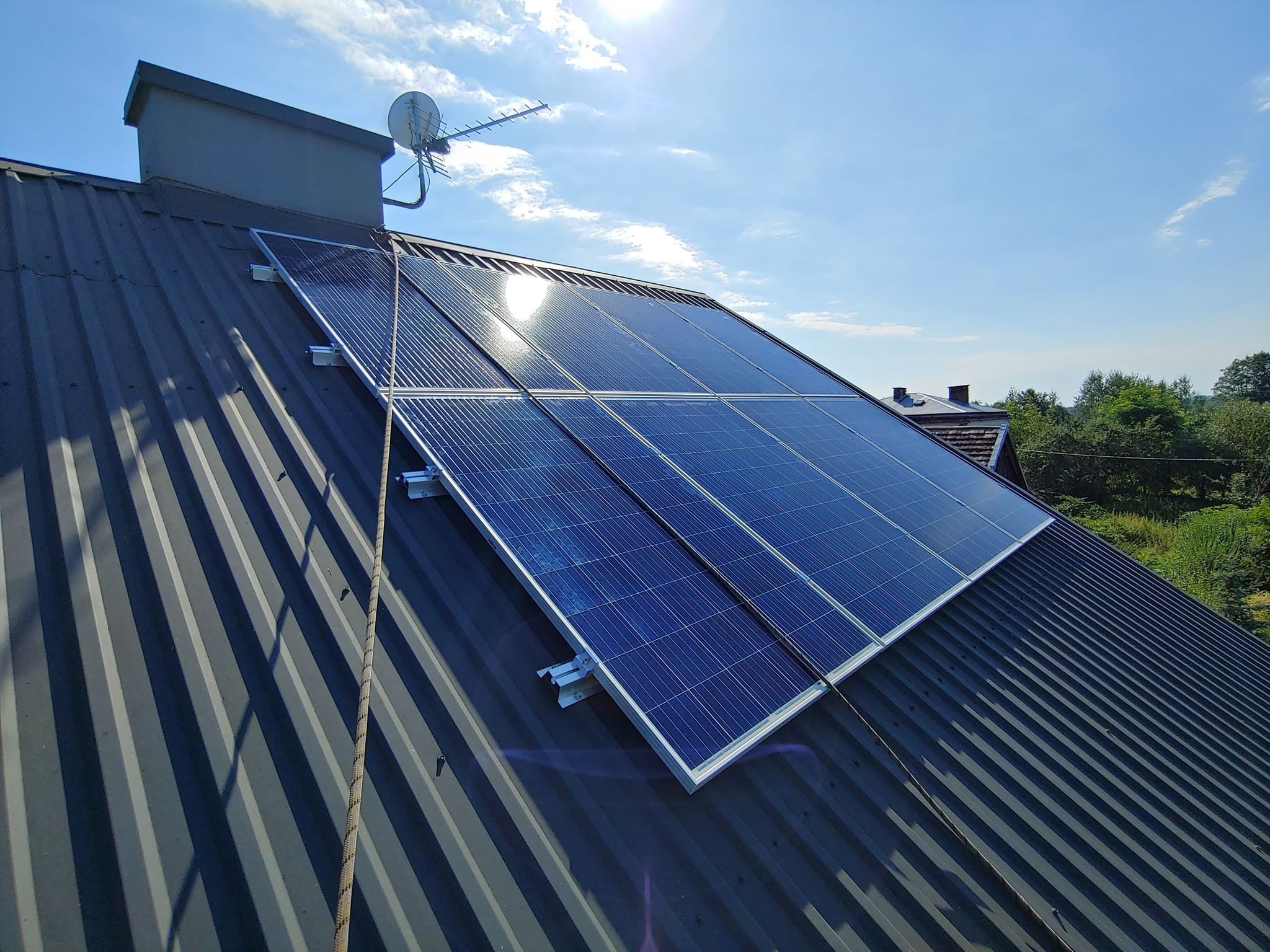Panel fotowoltaiczny zamontowany na dachu z blachy trapezowej, widoczna antena satelitarna i lina zabezpieczająca.