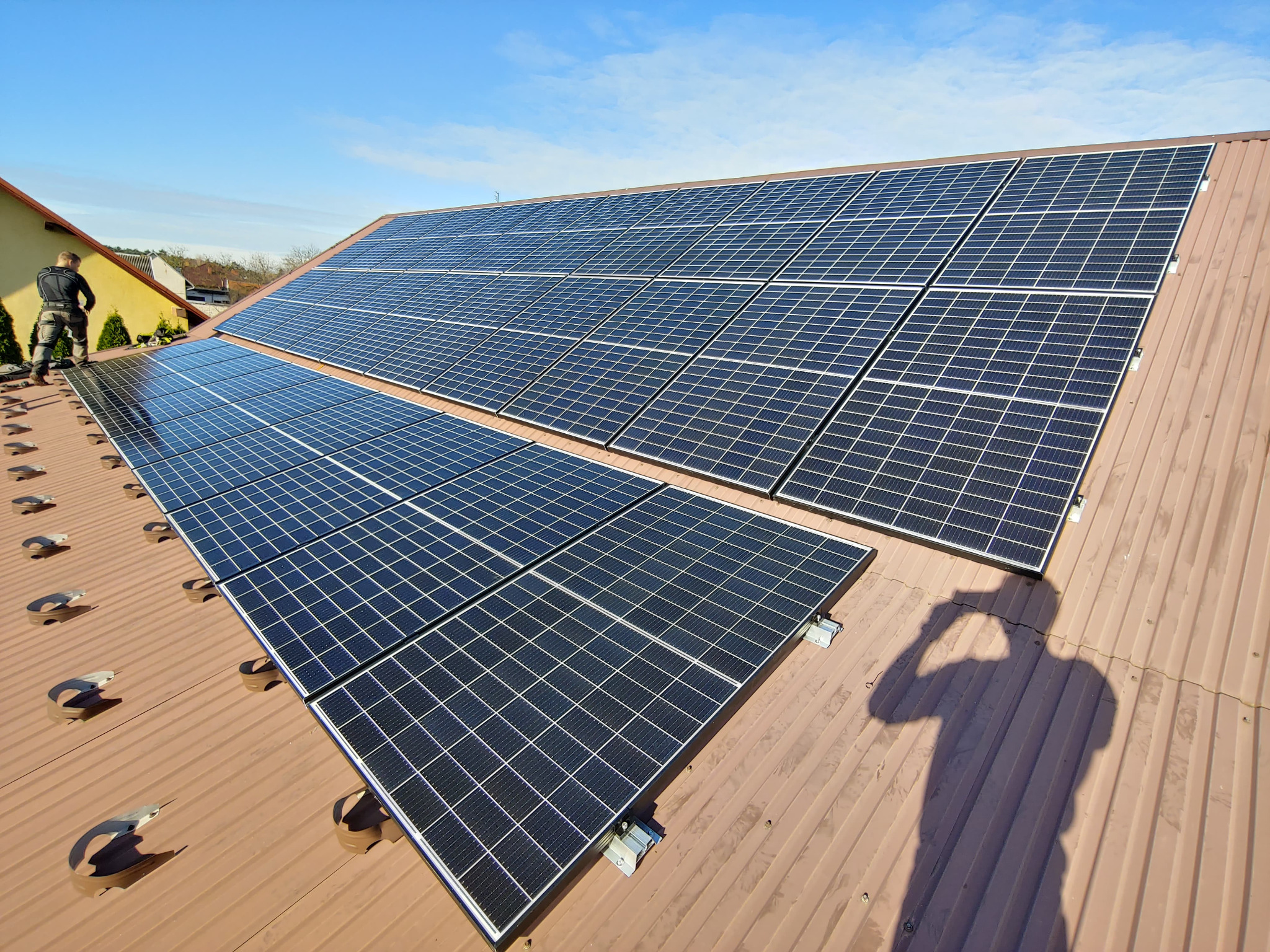 Instalacja paneli fotowoltaicznych na brązowym blaszanym dachu domu jednorodzinnego w słoneczny dzień, widoczny pracownik z narzędziami na dachu.