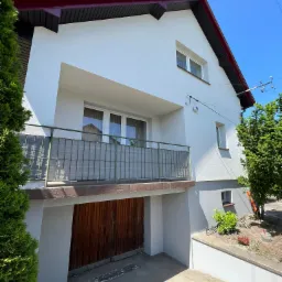 Dwupiętrowy dom z białymi ścianami, czerwonym dachem i drewnianą bramą garażową. Balustrada balkonu z szarą osłoną. Widoczny fragment ogrodu z roślinnością.