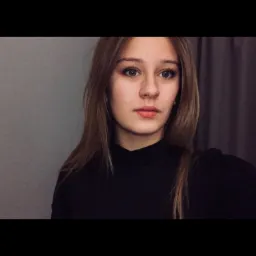 Portret młodej kobiety z długimi, prostymi włosami w czarnym golfie na tle szarej ściany i zasłony.