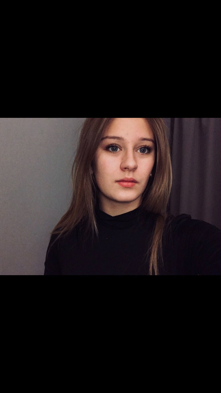 Portret młodej kobiety z długimi, prostymi włosami w czarnym golfie na tle szarej ściany i zasłony.